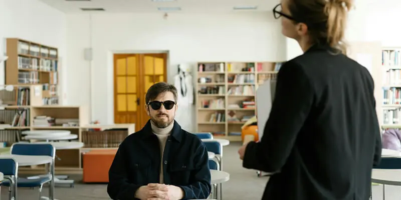 A blind man wearing dark sunglasses sits at a desk in a classroom setting, facing a person standing in the foreground with their back partially to the camera.