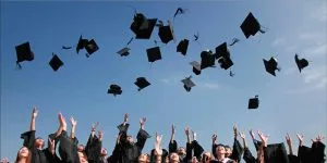 A group of graduates in black academic gowns joyfully tossing their caps into the air against a clear blue sky.