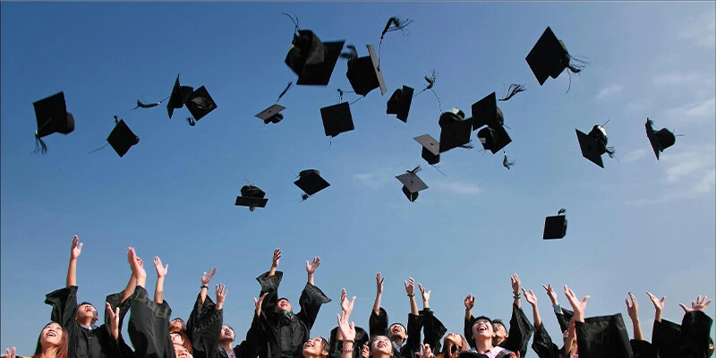 A group of graduates in black academic gowns joyfully tossing their caps into the air against a clear blue sky.