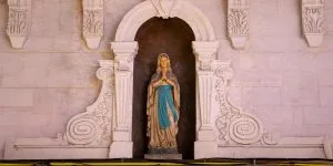 A statue of the Virgin Mary, with hands clasped in prayer, standing inside an ornate, carved stone alcove set into a wall.