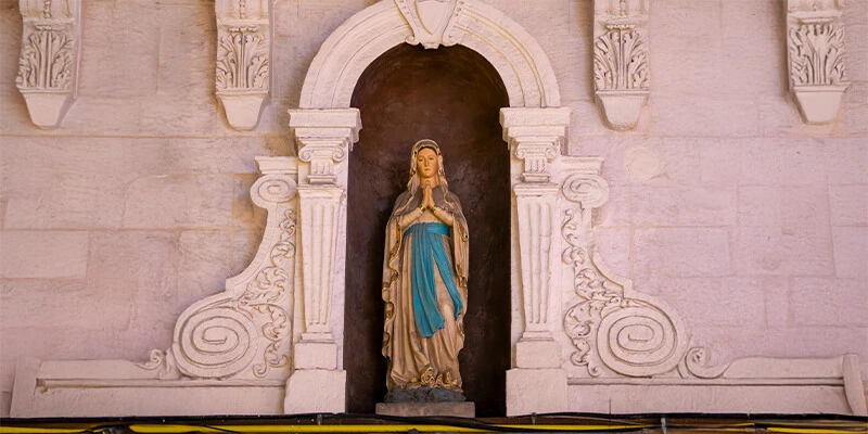 A statue of the Virgin Mary, with hands clasped in prayer, standing inside an ornate, carved stone alcove set into a wall.