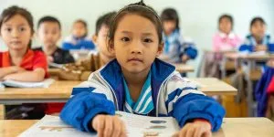 A young girl in a blue and white school jacket sits at a desk in a classroom, looking directly at the camera, with other students blurred in the background.