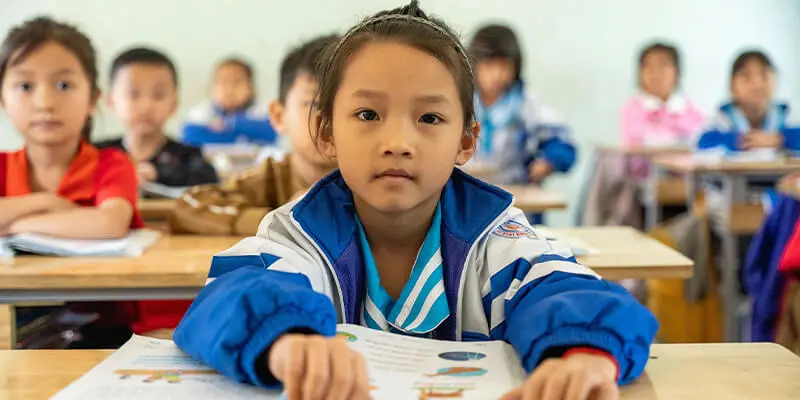 A young girl in a blue and white school jacket sits at a desk in a classroom, looking directly at the camera, with other students blurred in the background.