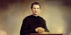 A portrait of Saint Don Bosco, depicted with a thin halo, wearing black clerical robes and standing behind a lectern with his hands clasped.