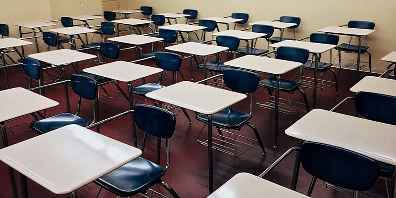 An empty classroom filled with rows of school desks, each with a white top and a blue chair, arranged on a red floor.