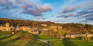 A wide aerial landscape shot of a sprawling campus of large, historic stone buildings nestled in a green valley at the base of a forested hill, illuminated by the warm light of sunrise or sunset under a partly cloudy sky.