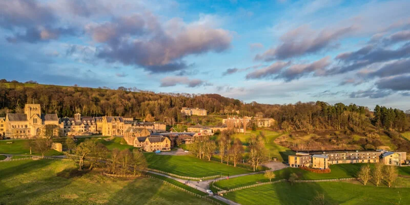 A wide aerial landscape shot of a sprawling campus of large, historic stone buildings nestled in a green valley at the base of a forested hill, illuminated by the warm light of sunrise or sunset under a partly cloudy sky.