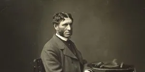 A vintage sepia-toned portrait of Matthew Arnold seated in profile, wearing a dark coat and sporting distinctive sideburns, with a table holding gloves visible beside him.