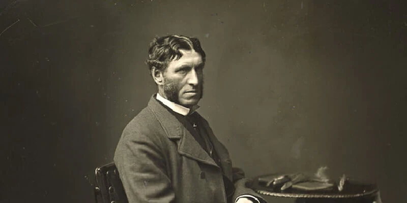A vintage sepia-toned portrait of Matthew Arnold seated in profile, wearing a dark coat and sporting distinctive sideburns, with a table holding gloves visible beside him.