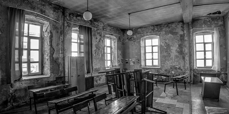A black and white photograph of a derelict, old-fashioned classroom with peeling walls, large windows, and rows of wooden desks, some of which are overturned.