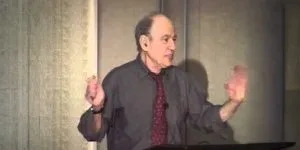 A photograph of Professor Barak Rosenshine giving a lecture, wearing a dark grey shirt and a red tie, gesturing with both hands raised as he speaks.