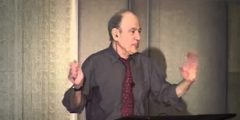 A photograph of Professor Barak Rosenshine giving a lecture, wearing a dark grey shirt and a red tie, gesturing with both hands raised as he speaks.