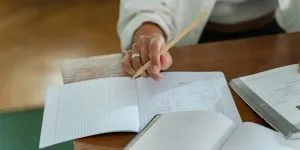 A close-up of a student's hand holding a pencil, poised to write in an open grid-paper notebook on a wooden desk during a study session.