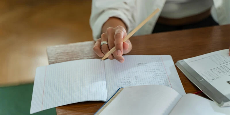 A close-up of a student's hand holding a pencil, poised to write in an open grid-paper notebook on a wooden desk during a study session.