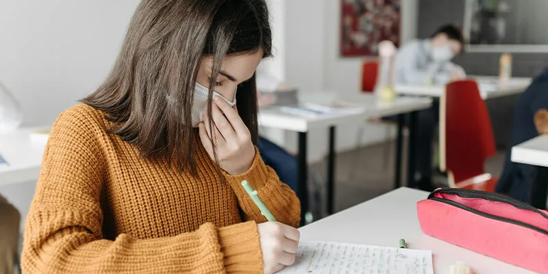 A young female student wearing a face mask and a brown sweater sits at a desk in a classroom, writing on a piece of paper.
