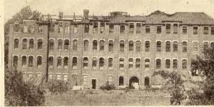 A historic, sepia-toned photograph of a large, multi-story brick building, the Ursuline school, appearing in ruins with a visibly damaged roof and many empty windows.